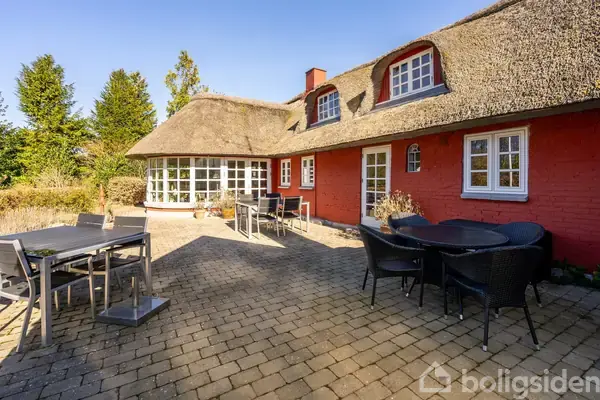 Stråtækt rødt hus med hvide sprossevinduer står stille i sollys, omgivet af en rummelig brolagt terrasse med sorte havemøbler. Grønne træer omkranser området.