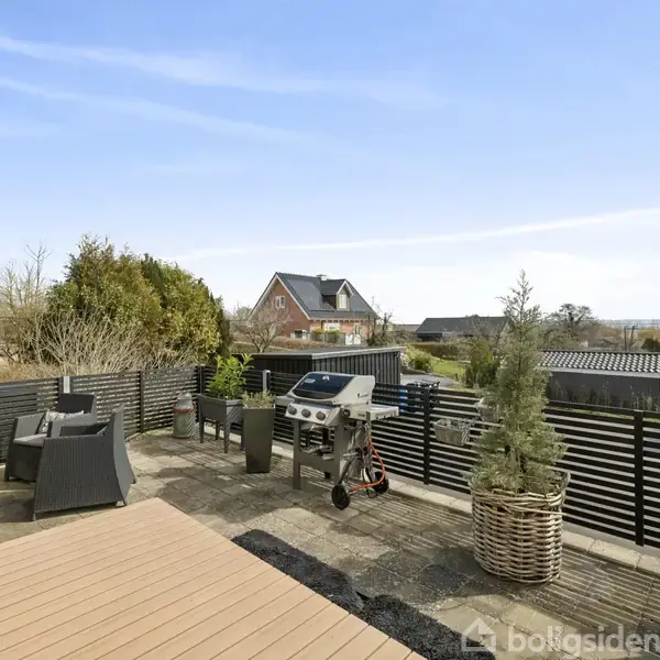 Terrasse med havemøbler og grill står stille i solbeskinnet haveomgivelse, omkranset af hegn og buske. I baggrunden ses huse og blå himmel, hvilket skaber en fredelig atmosfære.