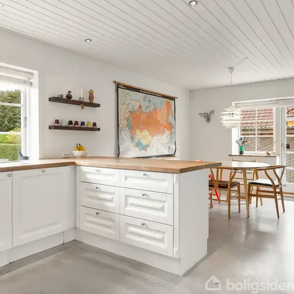 Et lyst køkken med hvide skabe og træbordplader har et vindue og udsigt til haven. En vægdekoreret verdenskort hænger over en reol. I baggrunden står et spisebord.