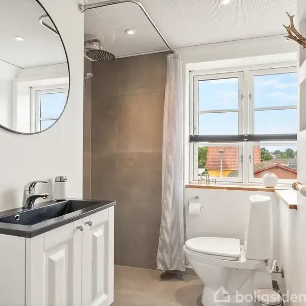 Et badeværelse med vask og spejl til venstre, og toilet til højre. Bruseområde bag hvidt forhæng ved vindue der viser udsigt til omgivelserne.