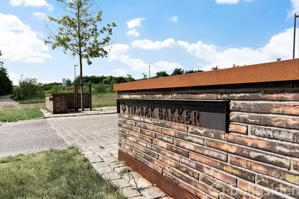En murstensmur med teksten "TULSTRUP BAKKER" står ved en vej omgivet af græs og træer under en blå himmel med skyer.