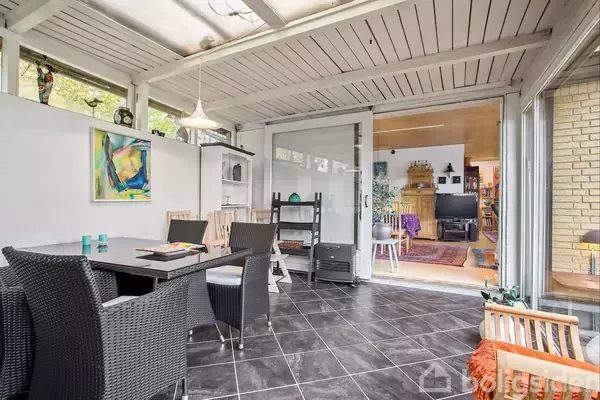 Et rummeligt udestueområde med sorte fliser og et bord med flettede stole. Glasdøre åbner til en varm, indendørs stue med hylder og sofaer, dekoreret i en jordnær stil.