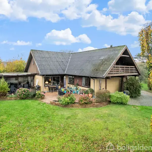 Hus med skråtag omgivet af frodig have; terrassen er udstyret med havemøbler. Træer og buske grænser op til græsplænen under en blå himmel med skyer.