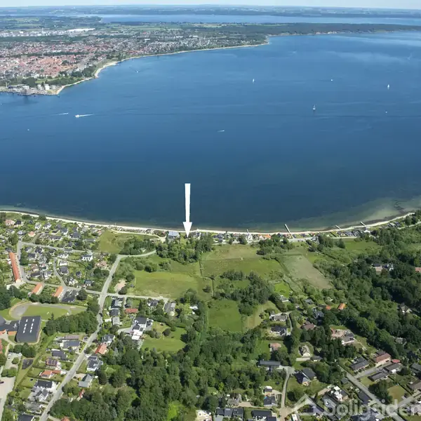 Kystlinje peget ud af pil omgives af grønne områder og by i forgrunden, mens det blå hav breder sig ud mod horisonten. Detaljer i beboelsesområdet ses tydeligt.