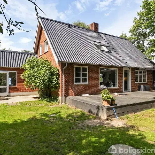 Rødt murstenshus med sort tag, omkranset af græsplæne og træer, har en træterrasse med en plantekrukke. En solrig have omgiver huset.