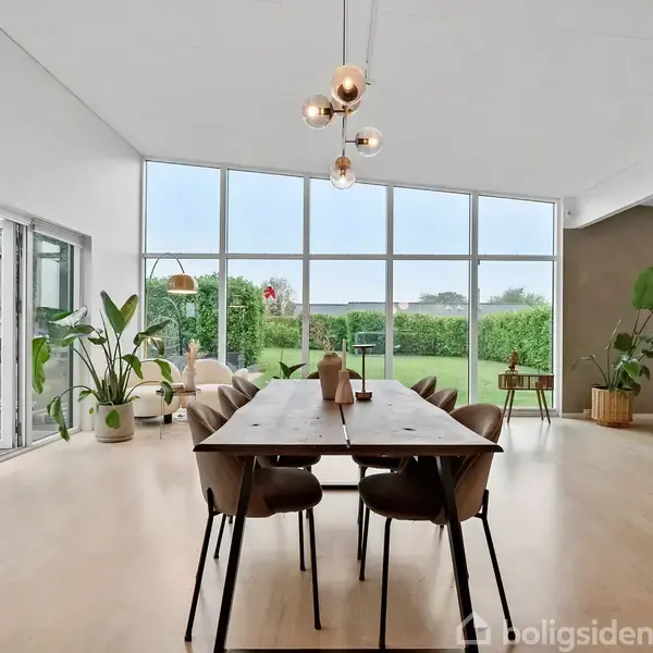Spisebord med stole står midt i et lyst rum med store vinduer, der vender ud mod en grøn have. Planter dekorerer hjørnerne, og en væg har et monteret TV.