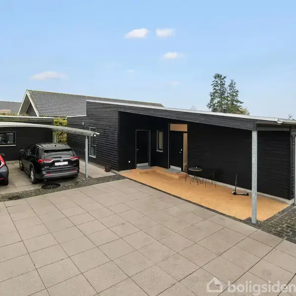 Sort, moderne hus med fladt tag og carport. Parkerede biler står i indkørslen. Foran huset er en lille terrasse med to stole. Træer og buske omgiver området.