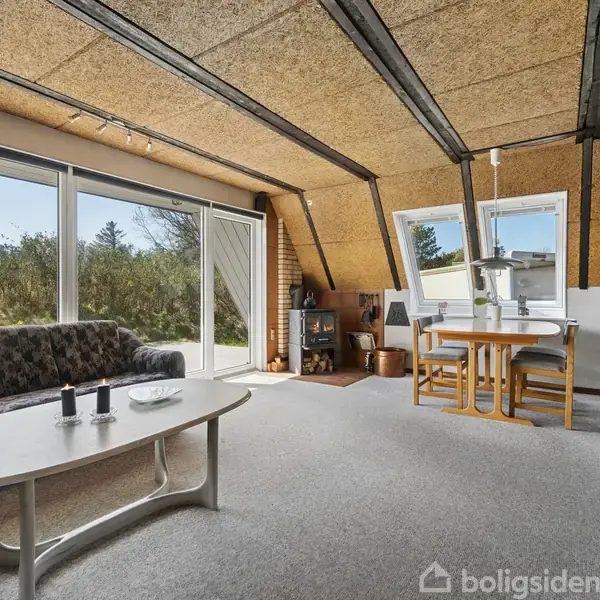 En stue med skrå lofter har en grå sofa ved siden af store vinduer. Et hvidt sofabord og en spiseplads ved vinduet. Udsigt til natur udenfor.