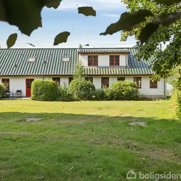 Hus med hvid facade og grønt tag står i en have med græs og buske. Der er også nogle træer, der danner en naturlig indramning.