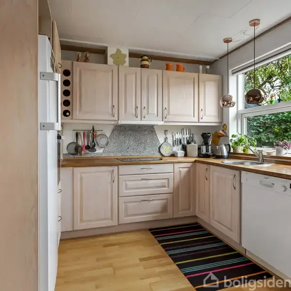 Et lyst køkken med træskabe og hvidevarer. På bordet står der køkkenredskaber, og et stort vindue fylder rummet med naturligt lys, udsigt til have. En stribet løber på gulvet.