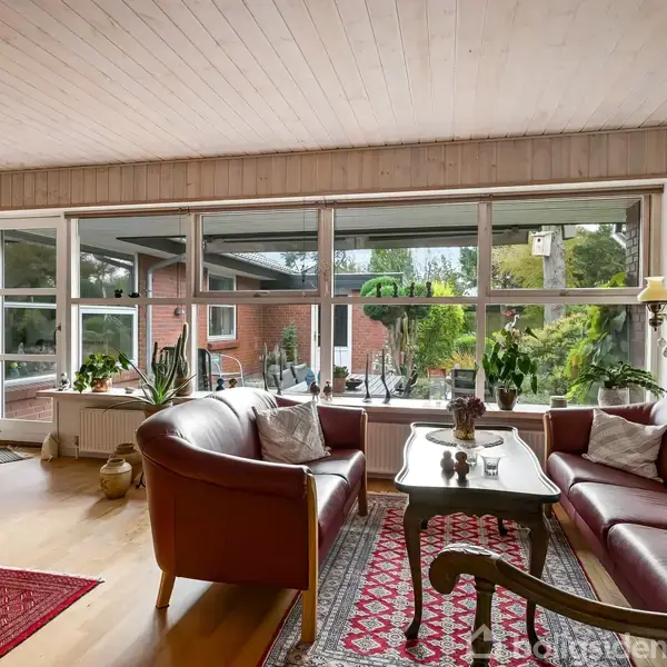 Et stueområde med røde lædersofaer og sofabord på orientalske tæpper. Store vinduer viser en have med buske og træer udenfor, og der hænger et maleri på væggen.