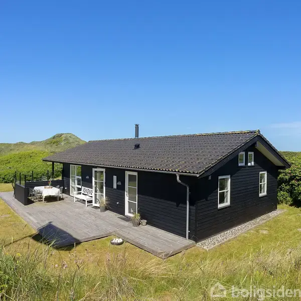 Et sort træhus med terrasse ligger i en frodig, grøn natur omgivet af bølgede bakker og under en klar blå himmel.