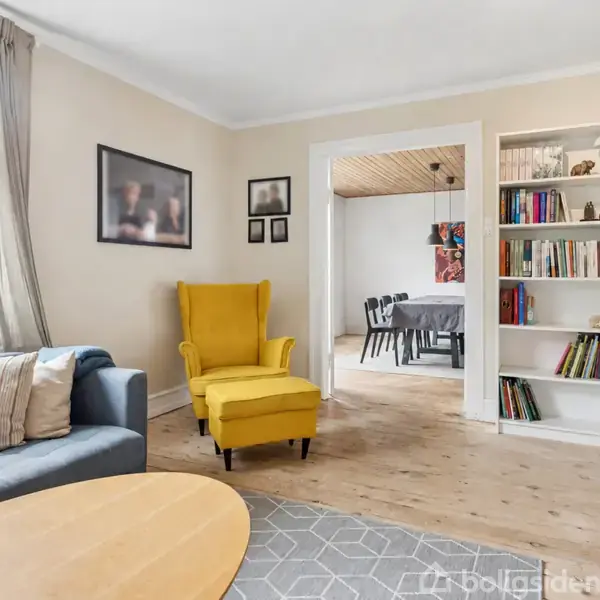 En gul lænestol står ved siden af en blå sofa i en stue med et trægulv og tæppe. I baggrunden ses en bogreol og et spisebord i en tilstødende spisestue.