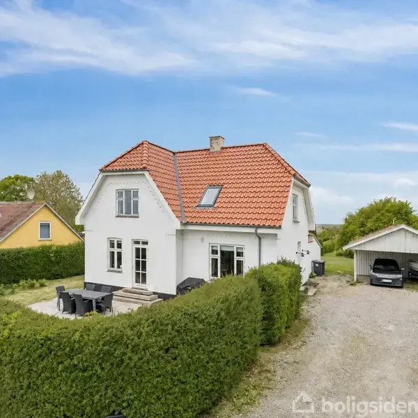 Hvidt hus med rødt tag står stille omgivet af en grøn hæk. I haven er et udendørs spisebord, og til højre er en grussti og en carport med bil.