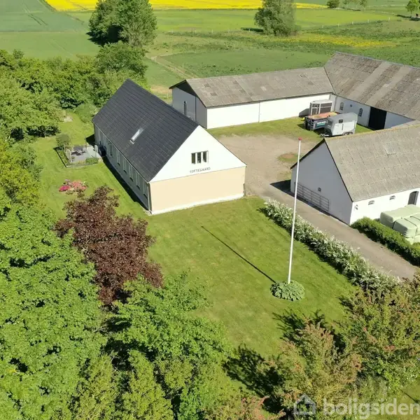 Gårdkompleks med to hovedbygninger omgivet af frodig, grøn natur. En flagstang står på græsset, og der er landbrugsland i baggrunden med marker og træer.