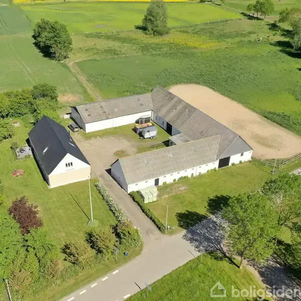 En firlænget gård omkranser en gårdsplads, beliggende i et landligt område med marker og træer. Der er flere bygninger med grå tage, omgivet af grønne marker.