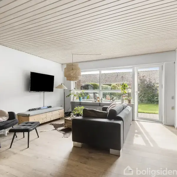 Stue med mørk sofa og lænestol, vægmonteret tv og træbord. Store vinduer giver udsigt til en grøn have. Trægulv og loft med lysindfald skaber en lys atmosfære.