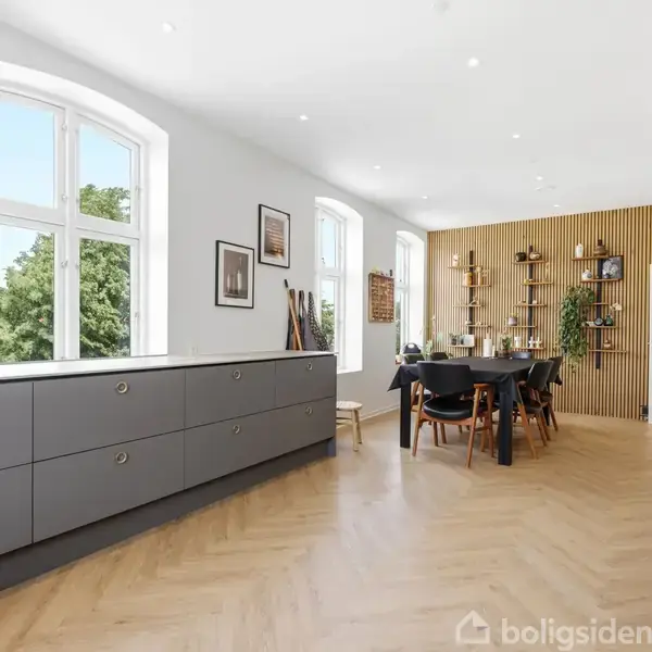 En lys spisestue med et stort, sort spisebord og stole, flankeret af moderne grå køkkenskabe. Store, buede vinduer giver udsigt til grønt træ udenfor. Elegant trægulv i sildebensmønster.