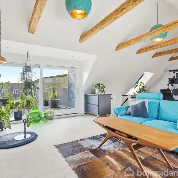 Lys stue med en turkis sofa og et træbord, omgivet af grønne planter. Store vinduer åbner ud til en altan. Synlige loftbjælker skaber en hyggelig atmosfære.