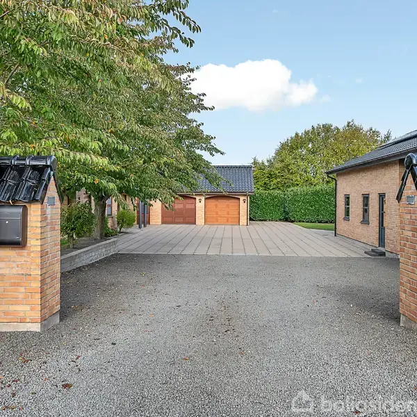 To murstenssøjler markerer indgangen til en gårdsplads med hæk på begge sider. En garage med brune døre ses for enden omgivet af grønne træer og græs.