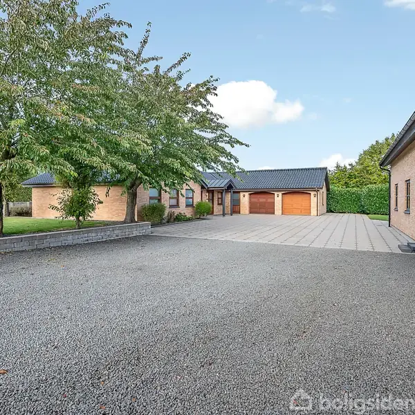 En række murstensbygninger omkranser en gårdsplads med grusbelægning. Til venstre ses træer og græsplæne, mens en klar, blå himmel danner baggrund.