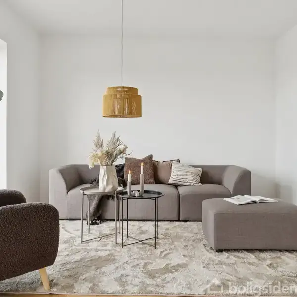 Grå sofa med puder står i stue nær brun lænestol og sofabord. Gul lampe hænger over bordene. Lyse vægge og tæppe skaber en lys, moderne atmosfære.