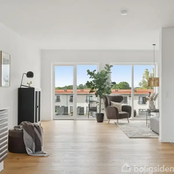 Stue med en stol og plante nær store vinduer, giver udsigt til en by. Et tæppe ligger på trægulvet. Vægge er hvidmalede, og der er minimalistisk indretning.