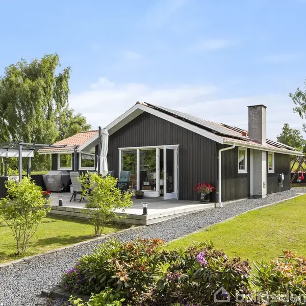 Sort hus med terrasse, omgivet af grønne græsplæner og blomsterbede, beliggende i en have med træer. En grussti fører til huset under en blå himmel.
