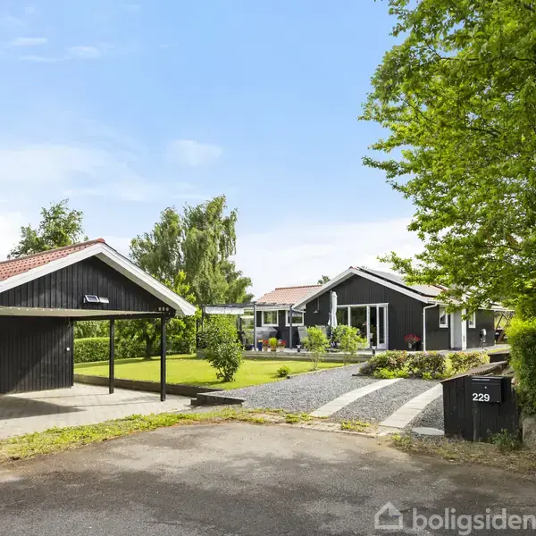 Carport ved et sortmalet hus med rødt tag. I forgrunden grusstier og græsplæne. Til højre en vej med skilt: "Til salg" og husetummer 229.