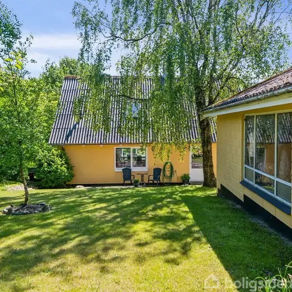 Et gult hus med skråt tag står i en have. Et træ kaster skygger over græsset, omgivet af frodig vegetation og en blå himmel.