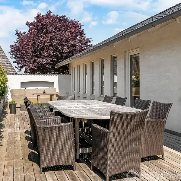 Et hvidt hus med gråt tag står centralt. En carport er ved siden af. Haven omkring har grønne buske og træer. Boligområdet har flere huse i baggrunden.