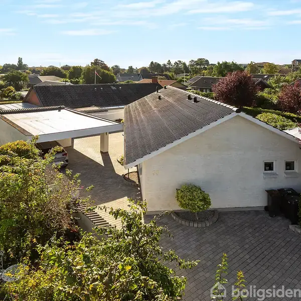 En hvid, moderne villa med sort tag ligger i et grønt boligområde. En terrasse med havemøbler og buske omkranser huset. En campingvogn er parkeret på indkørslen ved siden af.