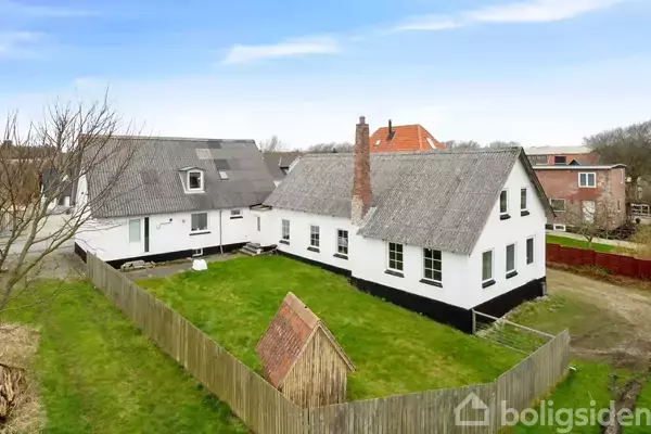 Hvide murstenshuse med stråtag ligger på en grøn græsplæne omgivet af et træhegn. Et lille skur står i forgrunden. I baggrunden ses flere huse i et boligområde.