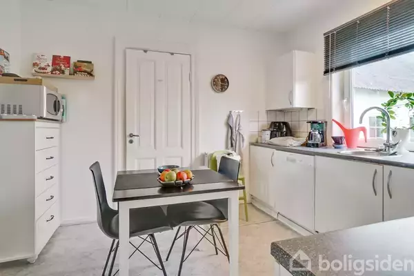 Et køkkenbord med frugtskål står i forgrunden. Omgivet af hvide skabe, mikrobølgeovn og vask med røde og sorte køkkenapparater. Et vindue lyser rummet op.