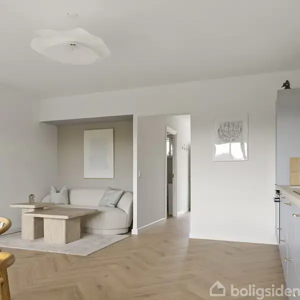 Stue med lyse møbler, hvor en sofa og sofabord er placeret i hjørnet. Til højre er et kompakts køkken. Rummets indretning er minimalistisk i et lyst og åbent rum.