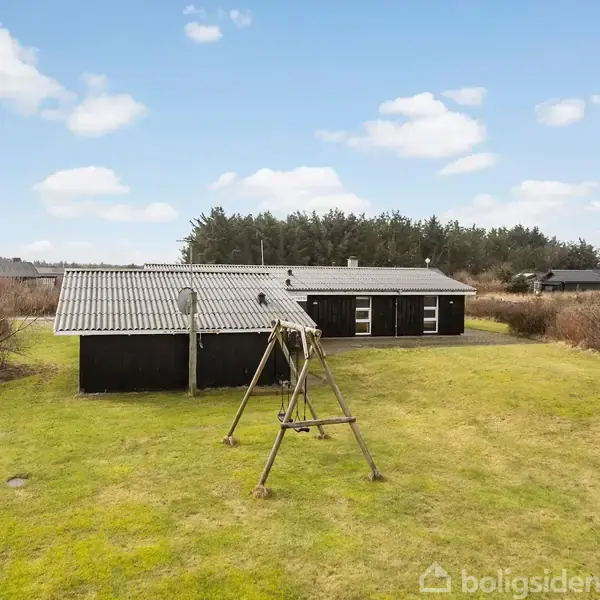 Et sort træhus står i en græsplæne med en gynge foran. Baggrunden er præget af skov og bygninger i det fjerne under en blå himmel med spredte skyer.