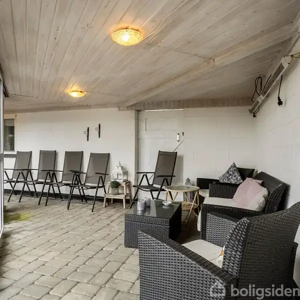 Terrassemøbler står på flisebelagt gulv. Flere stole står på række langs væggen. En lille sofa og to stole er placeret ved et sofabord. Væggene er hvide, og loftet er af træ.