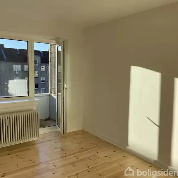 Et tomt rum har et åbent vindue og radiator. Sollys strømmer ind, og et stukket plankegulv ses. Udenfor er der en udsigt til naboens bygning.