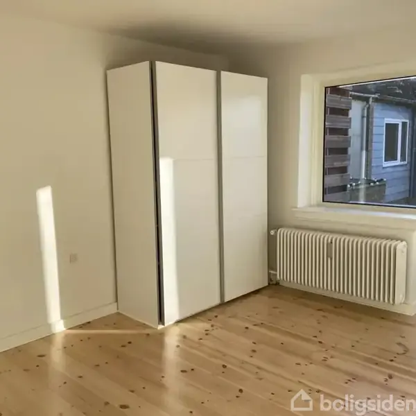Et hvidt garderobeskab står i et lyst rum med lakeret trægulv. Et vindue og en glasdør giver udsigt til nabobygninger. Radiator under vinduet.
