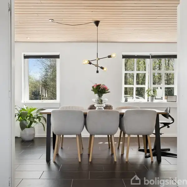 Spisebord med seks stole står i et lyst rum med store vinduer. En moderne lampe hænger over bordet, og der er en blomsterbuket på bordet. Planter og grøn udsigt ses udenfor.