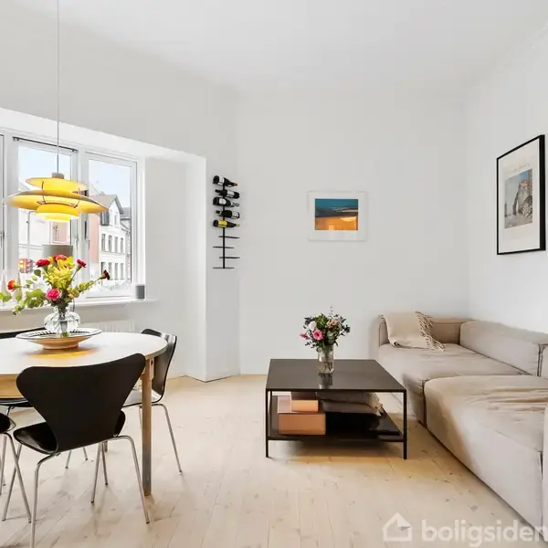 En lys stue med en beige sofa til højre, et sofabord med blomster og en rund spisebord med fire stole. Store vinduer giver udsigt til bygninger udenfor.