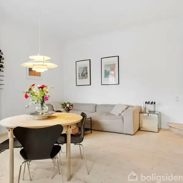 Et rundt spisebord med sorte stole står i en lyse stue. En moderne lampe hænger over bordet, og blomster pryder bordet. En sofa og billeder på væggen kompletterer indretningen.