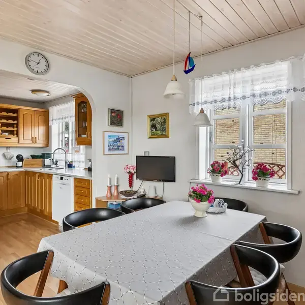 Et spisebord dækket med hvid dug omgivet af sorte stole. Baggrunden viser et køkken med træskabe og opvaskemaskine. Vindue med blomsterkasser og vægge dekoreret med billeder.