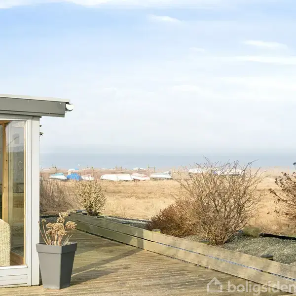 Glasveranda med hvide møbler står i en have med træterrasse; i baggrunden ses kystlinje og både på stranden.