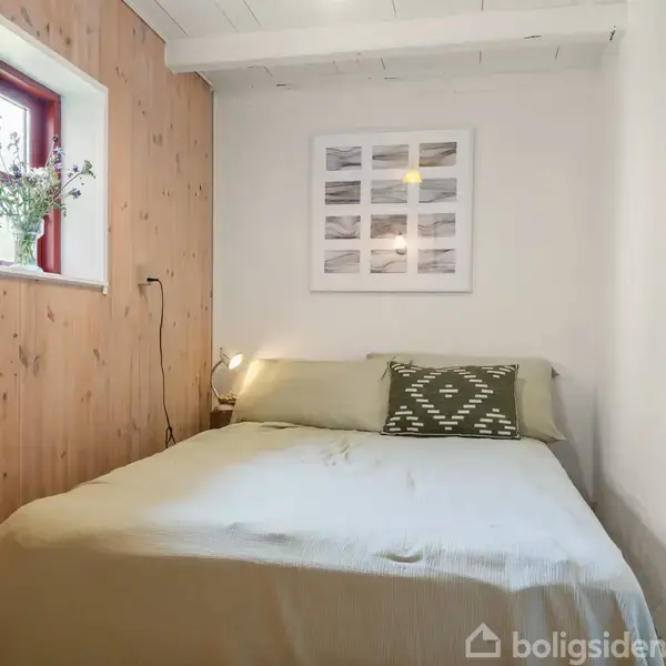 En seng står opredt med grønne puder i et lille rum. Ved siden af er der et vindue med planter og en lampe. Væggene er træ og hvidmalede.