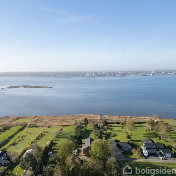 Kystlandskab med grønne marker og huse i forgrunden. Havet strækker sig gennem billedet, med en bylinje synlig i horisonten under en klar, blå himmel.