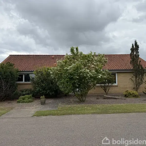 Et enkelt hus med rødt tegltag og murstensfacade er omkranset af forskellige buske og træer. Foran huset ses en grusgang, og himlen er overskyet.
