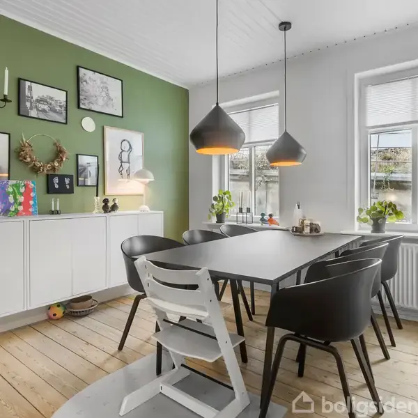 En spisestue med et sort bord omringet af sorte og hvide stole. Væggene har grønne og hvide flader. Der hænger billeder og dekorationer, samt planter, over en skænk.