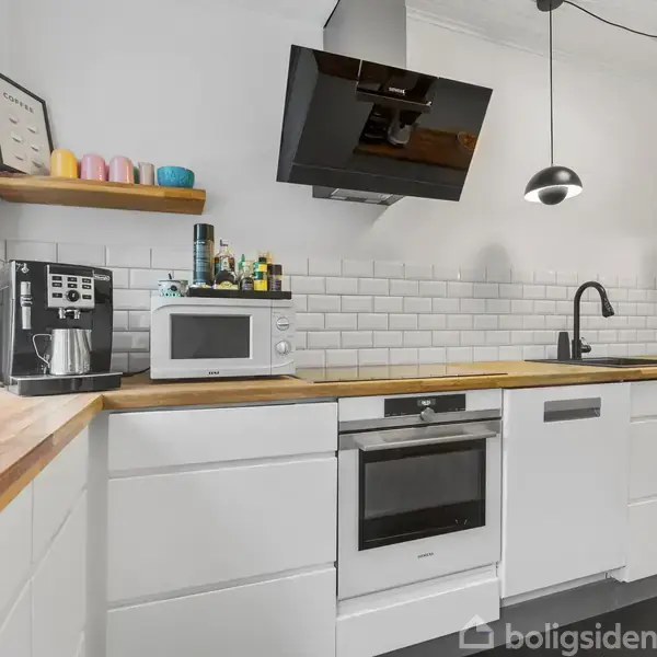 Et moderne køkken med hvidt interiør. Bordet har træoverflader, indbygget ovn, mikroovn og kaffemaskine. En sort emhætte hænger over komfuret. Væggene har hvide flisebelægninger.