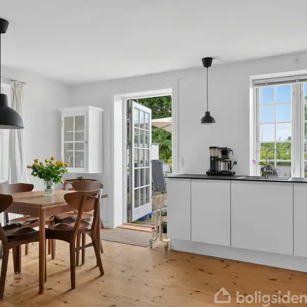 Træbord med stole står i køkken med hvide skabe og sorte lamper; vinduer åbne mod grøn have; dør fører ud til terrasse. Blomster pryder indretningen.